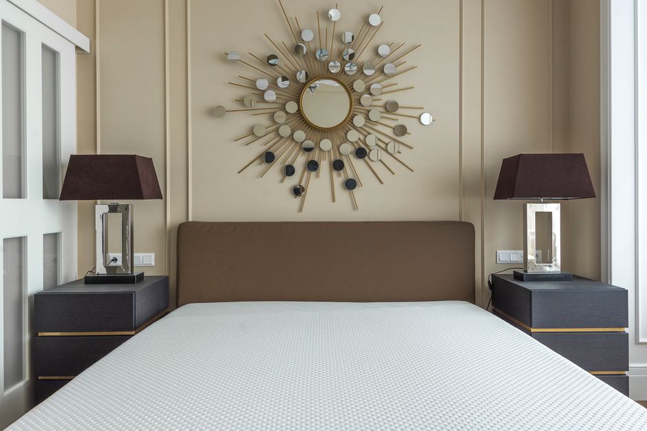 A contemporary bedroom featuring a large, neatly made bed with a beige upholstered headboard and a white textured bedspread. Flanking the bed are two matching dark-colored nightstands with sleek gold